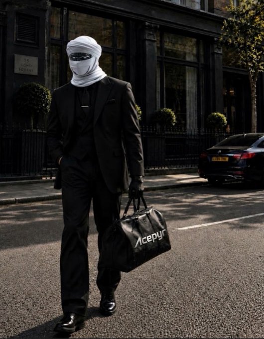 A person in a black suit walks down a street, their face wrapped in white bandages, carrying a black bag with the text 'Acepyr' printed on it.