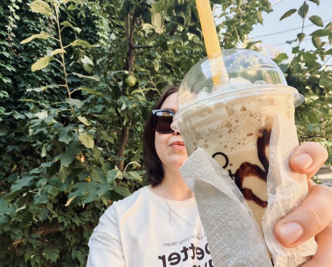 A woman wearing sunglasses and a white t-shirt holds up a clear plastic cup with a dome lid containing a brown and white layered beverage, possibly an iced coffee, with a straw and a napkin wrapped around it, set against green foliage.