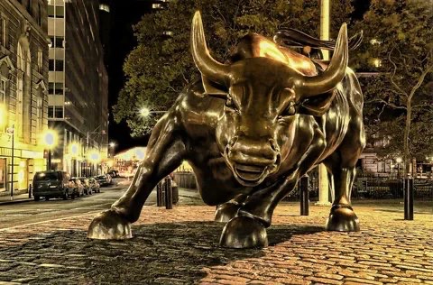 A bronze Charging Bull statue stands on a cobblestone street at night, illuminated by streetlights with a city street and buildings in the background.