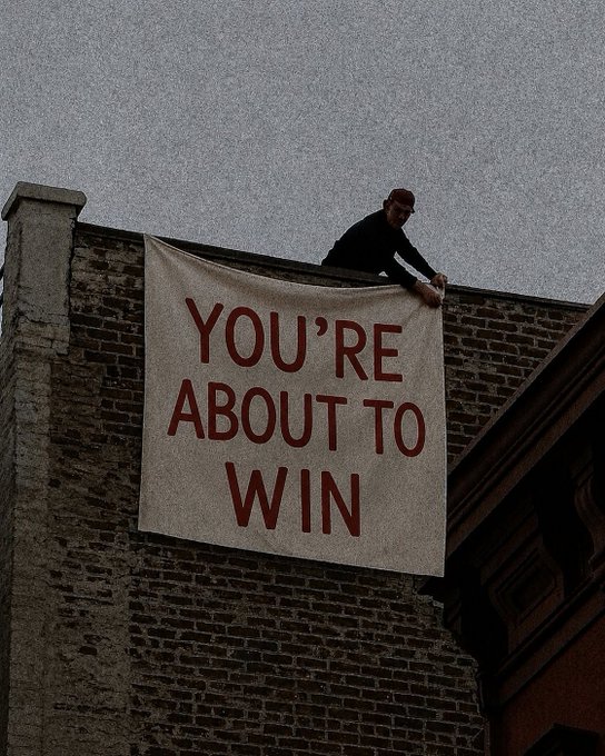 A person on a rooftop hangs a large white banner reading "YOU'RE ABOUT TO WIN" in red letters against the side of a brick building, with the "claim it!!!" caption reinforcing the bold, attention-grabbing message of imminent success.