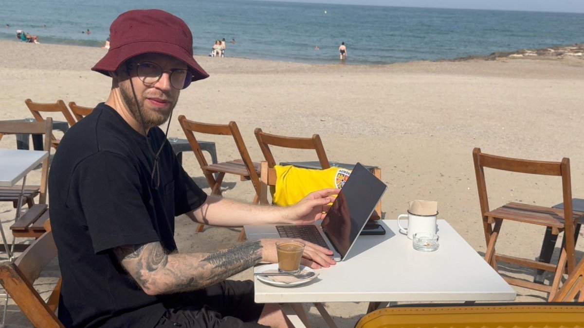 A man in a maroon bucket hat and glasses works on a laptop at a beachside café, with a yellow Spanish national team shirt draped over a chair beside him and the ocean in the background.