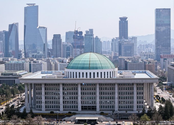 The image shows the National Assembly Building in Seoul, South Korea, with its distinctive green dome and classical colonnaded façade set against the city's modern skyline. The accompanying post discusses a 2026 parliamentary debate on cryptocurrency taxation policy, noting that starting January 1, 2027, virtual asset transfer and lending income will be taxed as "other income" at an effective rate of 22% after a 2.5 million won deduction, with the National Assembly serving as the legislative venue where these tax framework issues were addressed.