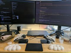The image shows a dual-monitor setup displaying lines of code, with a graphics tablet, phone, and other devices on a desk. It visually reinforces the caption's theme of "viral" tech-stack hacking—multiple codex accounts, forked codex, and self-hosted systems—being orchestrated from a compact, multi-device workstation, ironically framing the $33k/month token spend as the only thing being "burned."