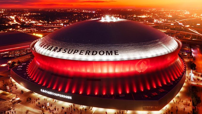 An aerial view of the Caesars Superdome illuminated in red at night with a sunset sky.
