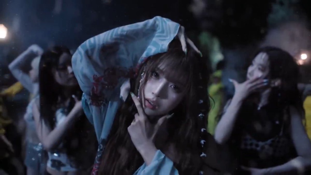 A close-up shot of a young woman with braided hair making a peace sign, accompanied by other women in a dimly lit setting.