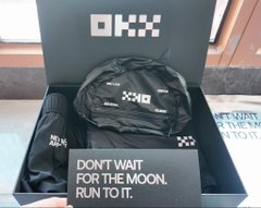 An open OKX merchandise box contains branded items including a 'DON'T WAIT FOR THE MOON. RUN TO IT.' promotional card.