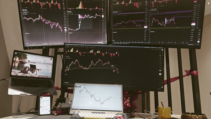 A multi-screen trading setup displays several stock charts across large monitors, with a laptop and smartphone also visible in the workstation arrangement.