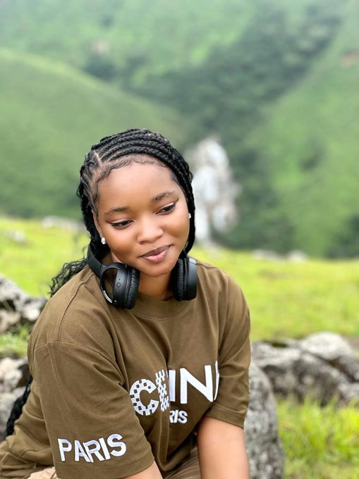 A woman with braided hair and headphones around her neck poses outdoors against a lush green mountainous backdrop, wearing an olive green t-shirt with "CHIN" and "PARIS" printed on it. The image captures a serene, reflective moment that complements the post's morning motivation to stay hydrated, engage with others, and share content.