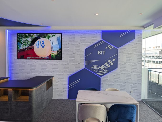 An interior view of a modern office space featuring hexagonal wall decorations labeled 'BIT', illuminated blue accent lighting, a wall-mounted TV screen, and a marble-topped conference table with blue chairs.