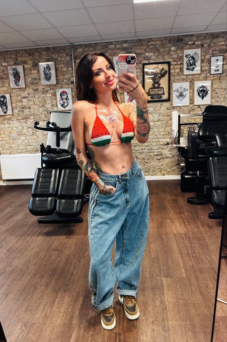 A tattooed woman taking a mirror selfie in what appears to be a tattoo parlor, wearing a watermelon-patterned bikini top, baggy jeans, and sneakers, with framed artwork on the brick wall behind her.