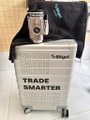 The image features a white suitcase with black text that reads "Trade Smarter" written on it, accompanied by a black bag or backpack placed next to it. The suitcase has wheels and handles for easy transportation. The background of the image shows a bathroom setting, which might suggest that someone is packing their belongings before traveling.
