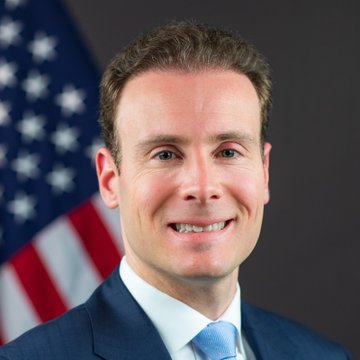 A smiling man in a dark blue suit with a light blue tie is photographed in front of an American flag. The image appears to be an official portrait of Michael Selig, identified in the accompanying post as CFTC Chair, who is discussing new cryptocurrency market regulations.