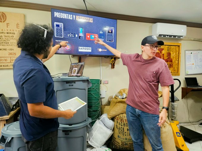 Two men stand in an indoor setting pointing at a wall-mounted television displaying a presentation titled "PREGUNTAS Y RESPUESTAS" that features various payment terminal devices and brand logos like "SATOSHI" and "ZBD".