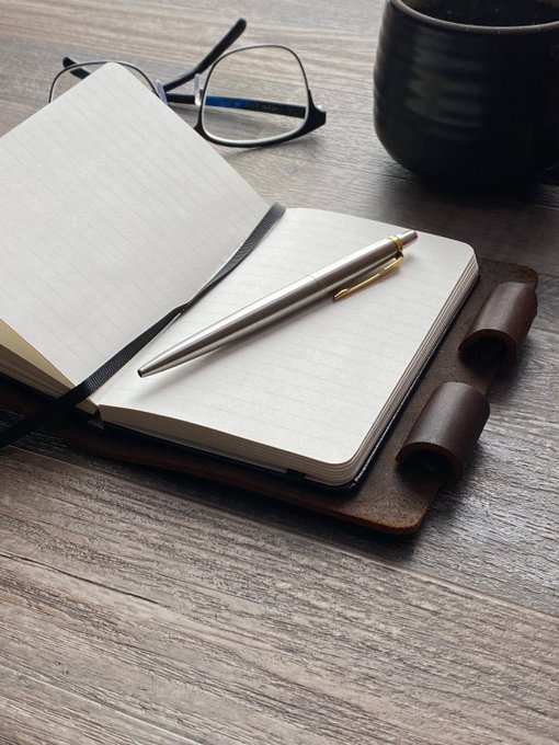 A flat lay photograph of an open blank notebook with a silver pen, a pair of black eyeglasses, and a dark ceramic mug on a wooden surface.