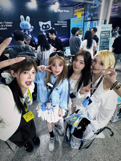 Four young women pose with peace signs at the Bitget Wallet booth, which displays its mascot characters and promotional banners reading "The Ultimate Wallet for Global Assets" and "✓Token #1 RWA Product," highlighting the crypto meetup where fans finally met Yoaka in person.