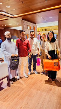 A group of five people, including one man in traditional white Arab attire, pose together indoors in what appears to be a shopping mall or hotel lobby, holding shopping bags.
