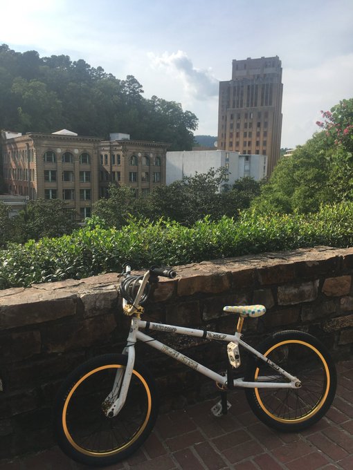 A white bicycle with yellow-rimmed wheels leans against a stone wall overlooking a cityscape with older brick buildings and a taller tan high-rise in the background.