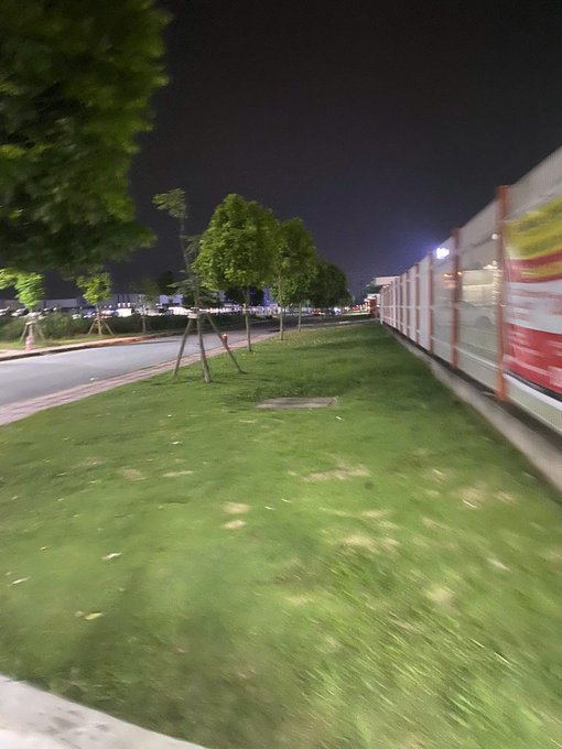 A blurry nighttime photo taken from a moving vehicle, showing a grassy strip lined with small trees, a road to the left, and a construction barrier to the right.
