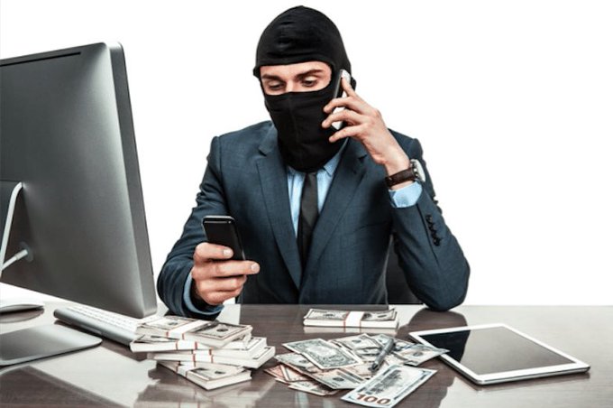 A masked man in a business suit uses a phone at a desk covered in stacks of hundred-dollar bills, with a computer monitor and tablet nearby.