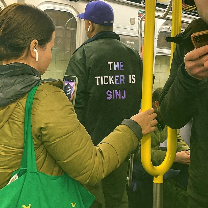 The image shows a woman holding onto a pole while riding on public transportation, possibly a subway car or bus. She has her arm around another passenger's shoulder, creating an atmosphere of camaraderie. The person she is holding onto is wearing a purple jacket with white text that says "The ticker is $SINJ". This message seems to be related to the cost of tickets for public transportation in the city.