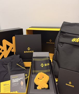 A Binance-branded 9YA anniversary gift box display featuring a "BINANCE" notebook, plush toy, book, and backpack, celebrating the exchange's 9-year anniversary.