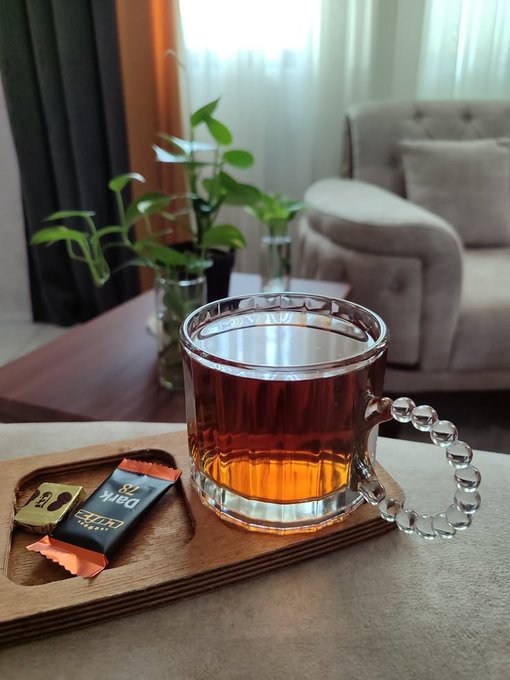 A clear glass mug filled with dark tea sits on a wooden tray alongside a wrapped snack and a small chocolate, with a potted plant and armchair in a softly lit room behind it.