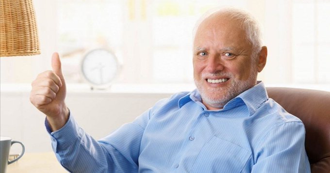 An elderly man in a blue button-up shirt smiles warmly while giving a thumbs-up gesture in a bright living room.