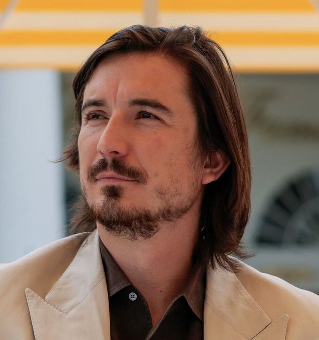 A man with shoulder-length brown hair, a beard and mustache, wearing a beige suit jacket over a dark shirt, gazes thoughtfully into the distance at what appears to be an outdoor event under a yellow canopy. The image contrasts the Breaking news of Robinhood's chain fees surpassing those of Solana and BNB Chain, perhaps symbolizing the moment of "shock" or awe at the unexpected rise of the newcomer platform in the crypto space.
