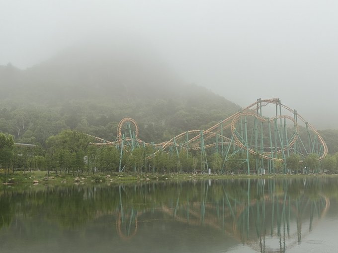 A roller coaster track, painted green and orange, curves through a foggy landscape with trees and mountains in the background.