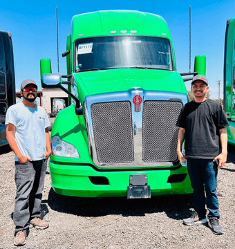Two men stand on either side of a bright green Kenworth semi-truck, with the number '2117' visible on the windshield.