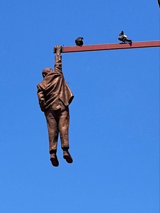 The image features a statue of a man hanging from a pole, with another bird perched on his shoulder. The statue depicts the man as wearing a suit and tie, while the bird has its wings spread out. The scene appears to be set against a clear blue sky, creating an interesting contrast between the man's attire and the bird's natural plumage. The post accompanying this image reads "Back again", which could potentially be a reference to the statue or the act of hanging from the pole.