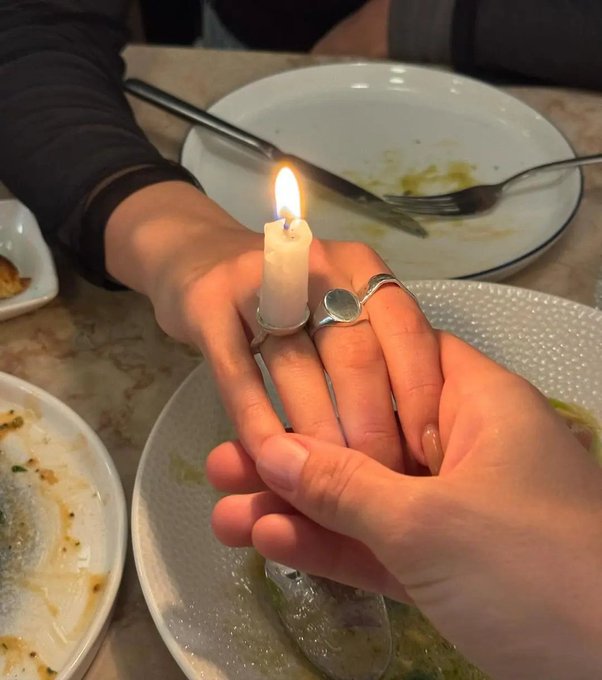 Two hands gently hold a lit white candle on a dining table surrounded by empty plates and silverware, with a silver ring visible on one finger.