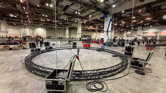 A large empty convention hall features a circular metal truss suspended by chains, surrounded by equipment cases and rigging gear.