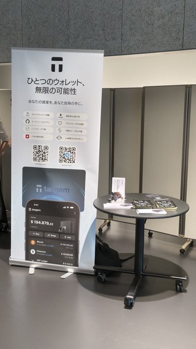 A promotional banner for the Tangem crypto hardware wallet with Japanese text reading 'ひとつのウォレット、無限の可能性', displayed at a booth with a mockup of the wallet showing a balance of $104,879.42.