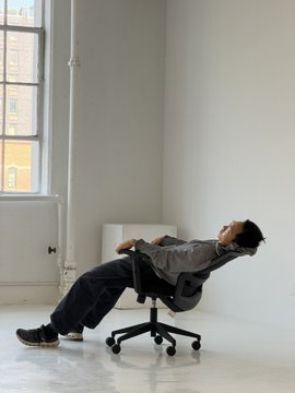 A man leans back dramatically in an ergonomic office chair in a minimalist white room with large windows, showcasing the SideShift V3 chair during its launch video shoot.