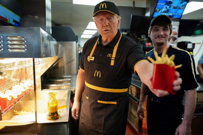 We need to write a concise 1-2 sentence caption describing what is in the image, including any key text, numbers, tickers or names visible. Also note how the image relates to the post.

Image: A person resembling Donald Trump (actually a lookalike) wearing a black McDonald's uniform with yellow accents, cap with "M" logo, apron with "M". He is holding out a red container of fries. Another person in similar uniform stands behind. In background, kitchen equipment, a screen showing maybe "M". There's a name tag "Trump" maybe? Actually the name tag says "Trump". Let's look: The name tag on his apron appears to say "Trump". Yes, there is a name tag on the left side of his apron that reads "Trump". So key text: "M" logo on cap and apron, "Trump" name tag. Also the background screens maybe show "