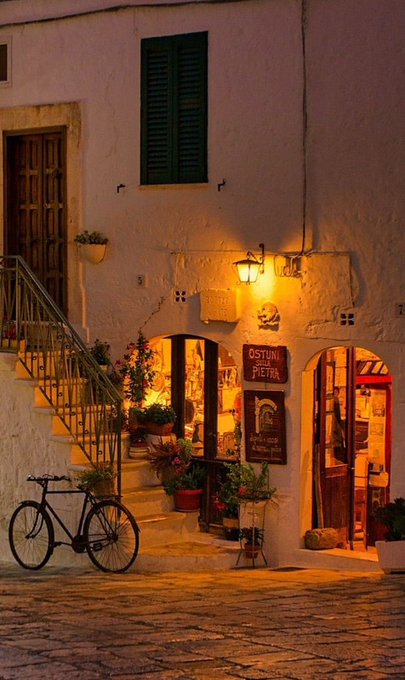 An evening scene of a charming cobblestone street in what appears to be Ostuni, Italy, featuring illuminated stone archways, potted flowers, a parked bicycle, and a sign reading 'OSTUNI IN PIETRA'.