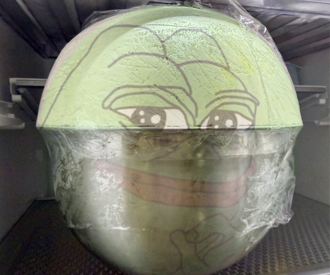 A large, round, green object wrapped in clear plastic featuring a crudely drawn face with furrowed brows, a mustache, and an angry expression, sitting on a metal surface.