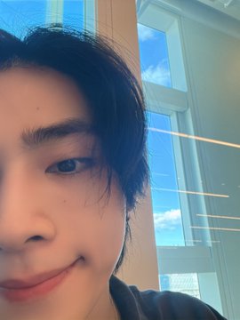 A close-up selfie of a young person with dark hair against a bright window with a blue sky view.