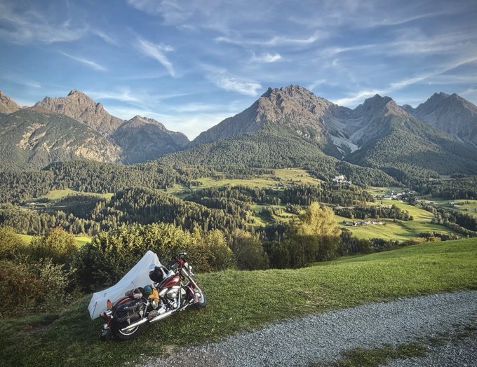 A motorcycle parked on grass at the edge of a gravel path overlooking a stunning mountain valley with green meadows, dense forests, and rugged peaks under a partly cloudy sky.