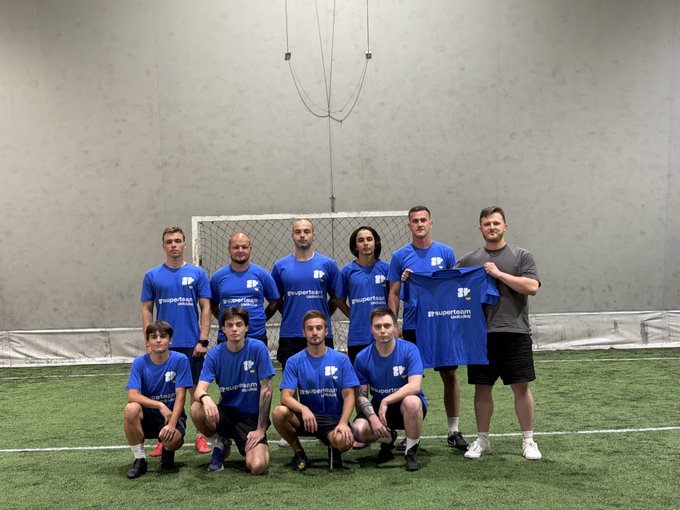 A sports team in matching blue Superteam jerseys poses for a group photo on an indoor soccer field, with one player holding up an extra jersey.
