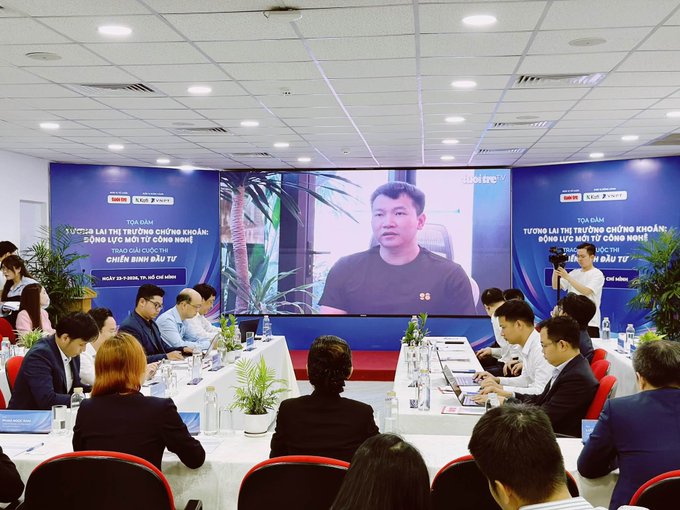 The image shows a business conference where a man is being featured on a large screen, with banners displaying "TOẠ ĐÀM VỀ LÀM THẾ NÀO ĐỂ ỨNG DỤNG BLOCKCHAIN VÀO ĐỜI SỐNG TỪ CÔNG NGHỆ" (a discussion on how to apply blockchain technology to daily life) and "ĐẦU TƯ" (Investment). The setup, featuring a prominent display of the speaker, aligns with the post's reference to a prominent figure in the crypto/KOL community (likely anh Vinh of Coin98) addressing attendees at what appears to be a blockchain industry event.
