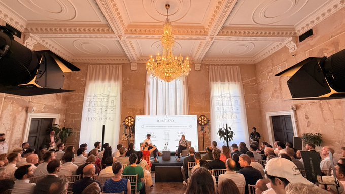 An elegant indoor event space with a chandelier, ornate ceiling, and an audience seated watching a presentation on a screen.