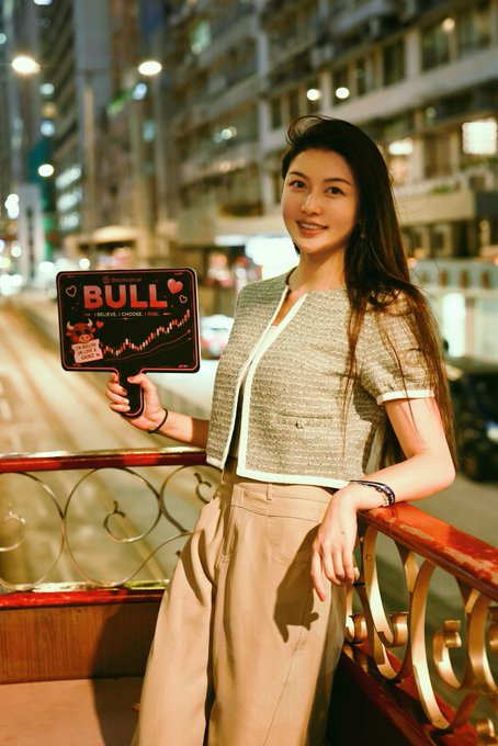 A woman stands on a city balcony at night holding a sign that reads "BULL - I BELIEVE I HAVE FEELINGS," set against a backdrop of illuminated high-rise buildings.