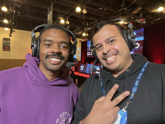Two smiling men wearing headsets posing together at what appears to be a gaming or tech convention, one in a purple hoodie and the other in a black hoodie making a peace sign.