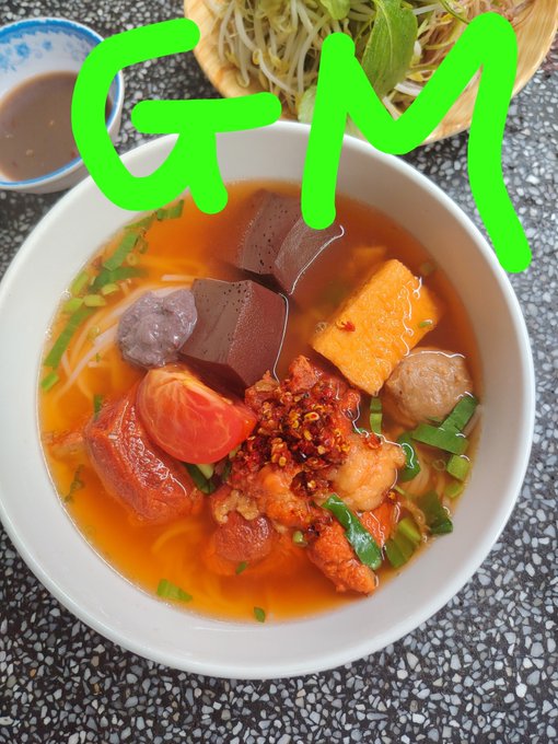 A bowl of Vietnamese soup (bún riêu) with crab, tomato, blood tofu, and noodles, labeled 'G-M' in large green handwritten text overlaid on the image.