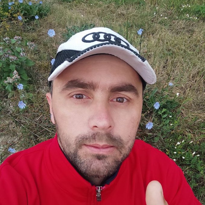 A selfie of a man wearing a white Audi cap and red jacket, standing in a field with wildflowers, giving a thumbs up.