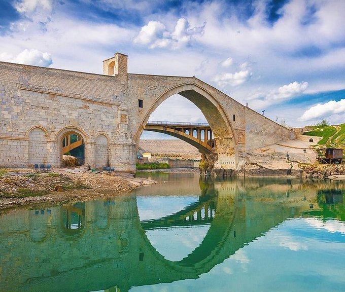 A photograph of an ancient stone arch bridge spanning a calm, turquoise river under a blue sky with scattered clouds.