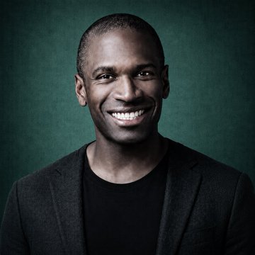 A professional headshot portrait of a smiling Black man with a shaved head wearing a black blazer over a black t-shirt, set against a dark green textured background.