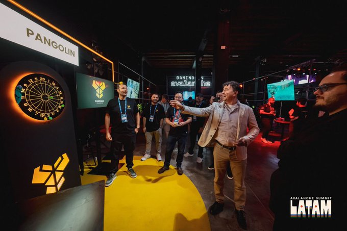 A man throws a dart at a Pangolin-branded dartboard while others watch at Avalanche Summit LATAM, capturing a nostalgic event moment as Pangolin invites connections to reunite at the upcoming NYC summit.